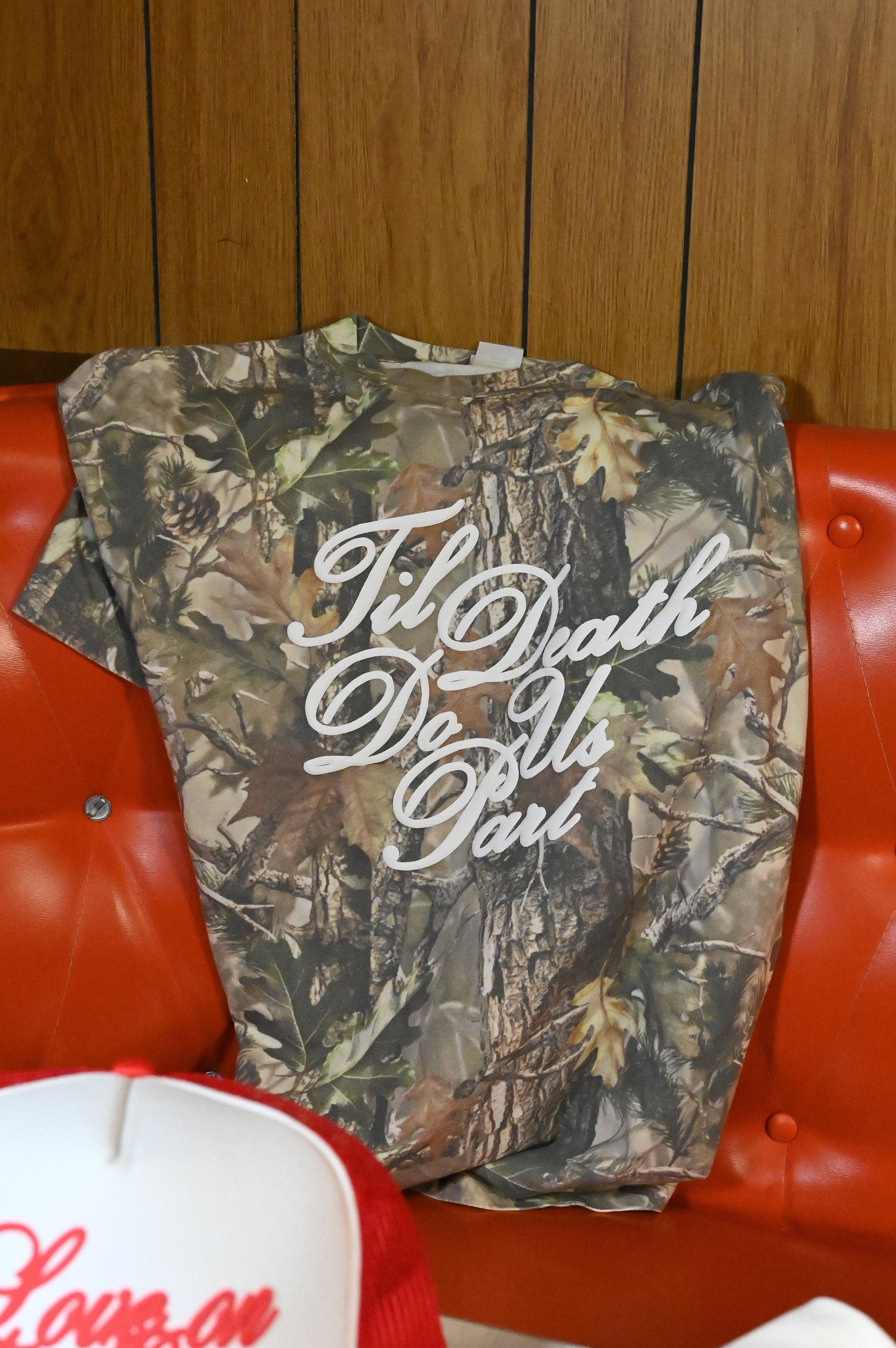 Camouflage shirt with text, red cap with white brim and text, and white shirt with blue text on a red couch.